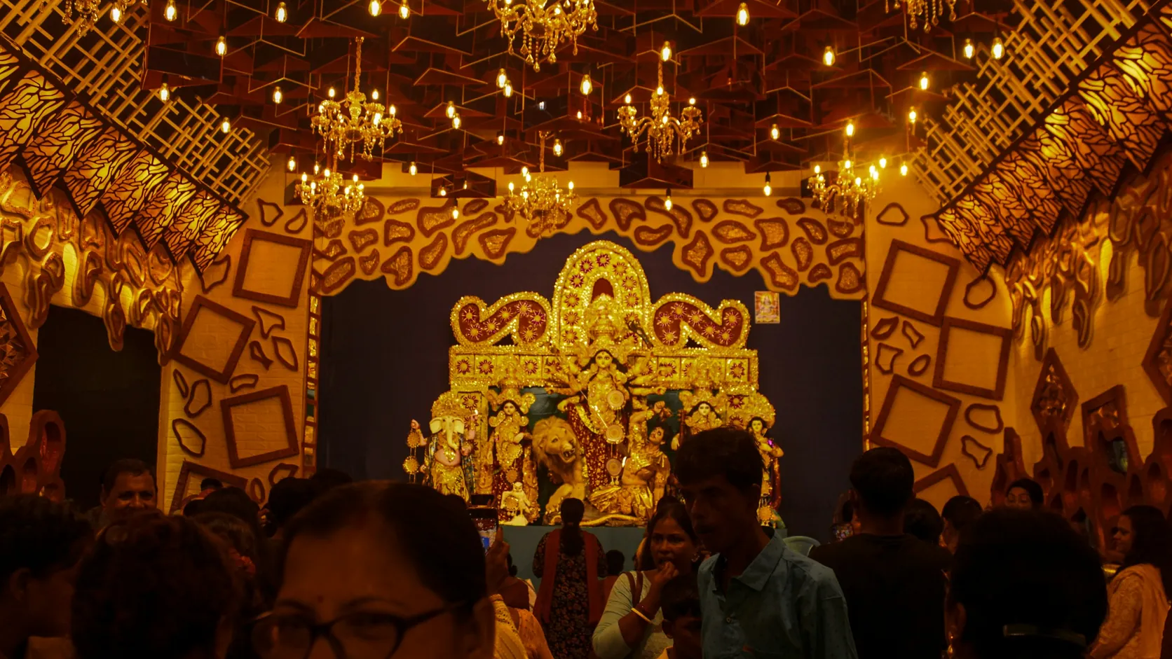 An evening crowd under chandeliers inside a gold-decorated Durga Puja pandal in Kolkata, with the idol at the far end
