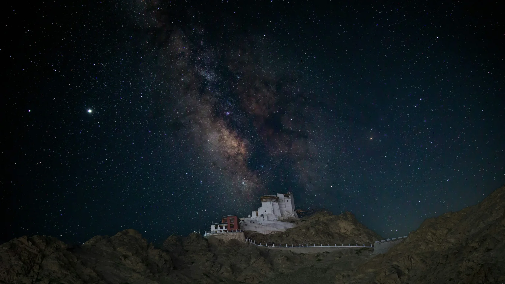 The Milky Way arching over a small lit monastery on a dark ridge in Ladakh