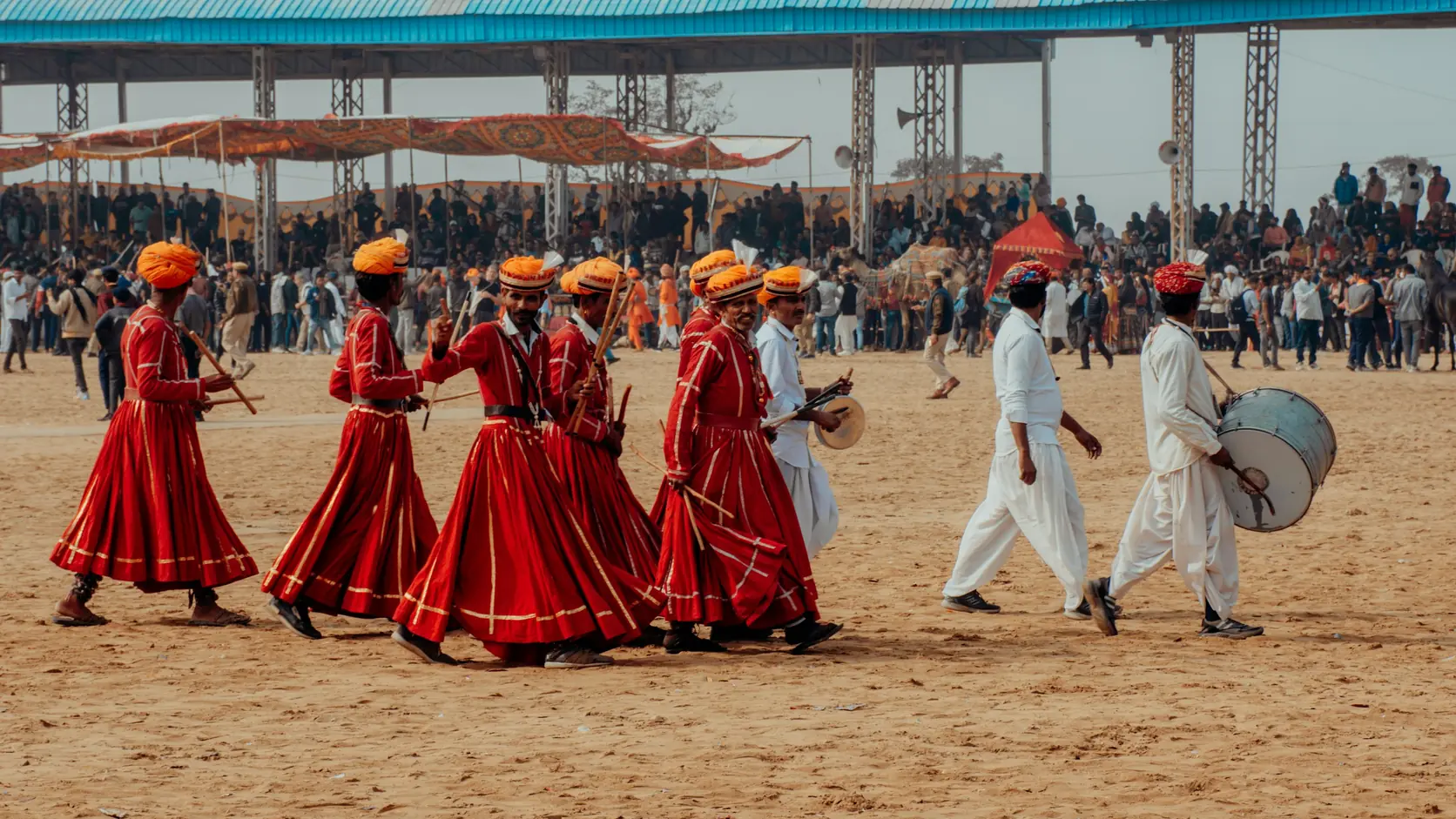 Folk dancers in red robes and orange turbans with a drummer, performing on the sand of the Pushkar fair arena in front of a crowd