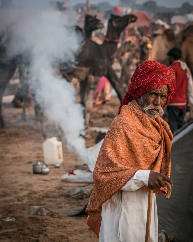 An elderly herder in a red turban and orange shawl among camels and morning smoke at the Pushkar fairground